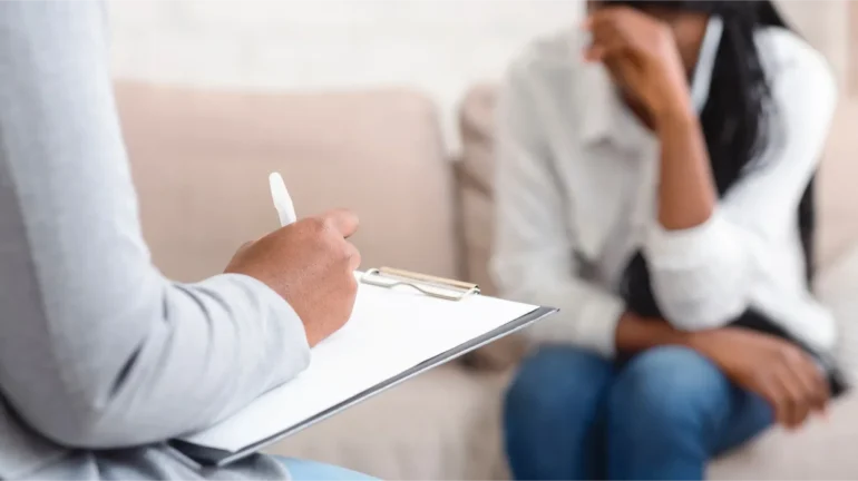 Photo of seated psychiatrist (from the back, right arm, writing on a pan in her lap) and a depressed woman sitting on a light sofa across from her, indistinct