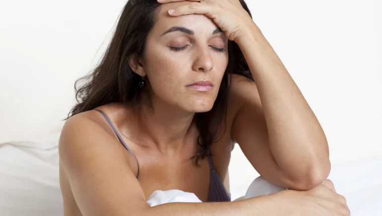 Photo of a woman sitting on a bed, eyes closed, depressed expression, hugging her knees with one arm, the other elbow on knee, hand on her brow