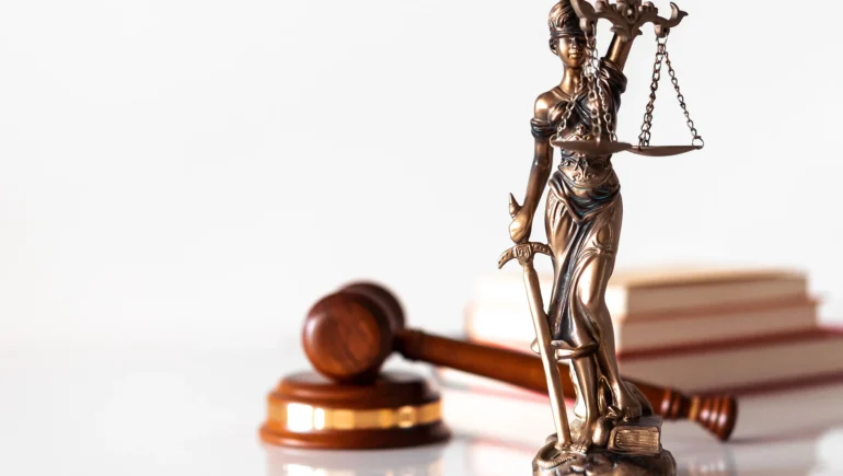 Photo of a bronze statue of goddess of justice holding up a scale and a sword in the other hand, behind her a judge's hammer and some books on a white surface